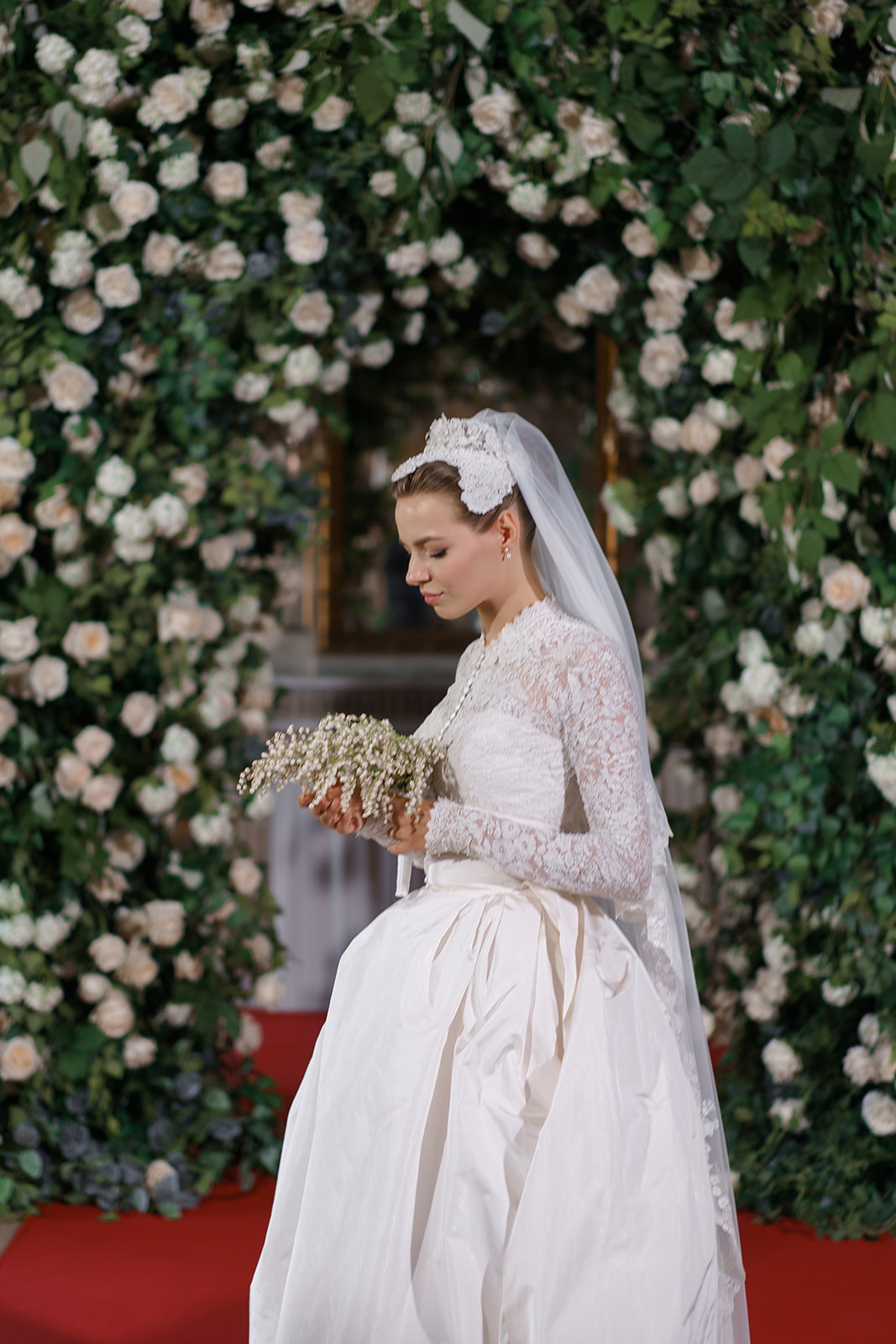Grace Kelly inspired bride poses with veil in front of white rose arch
